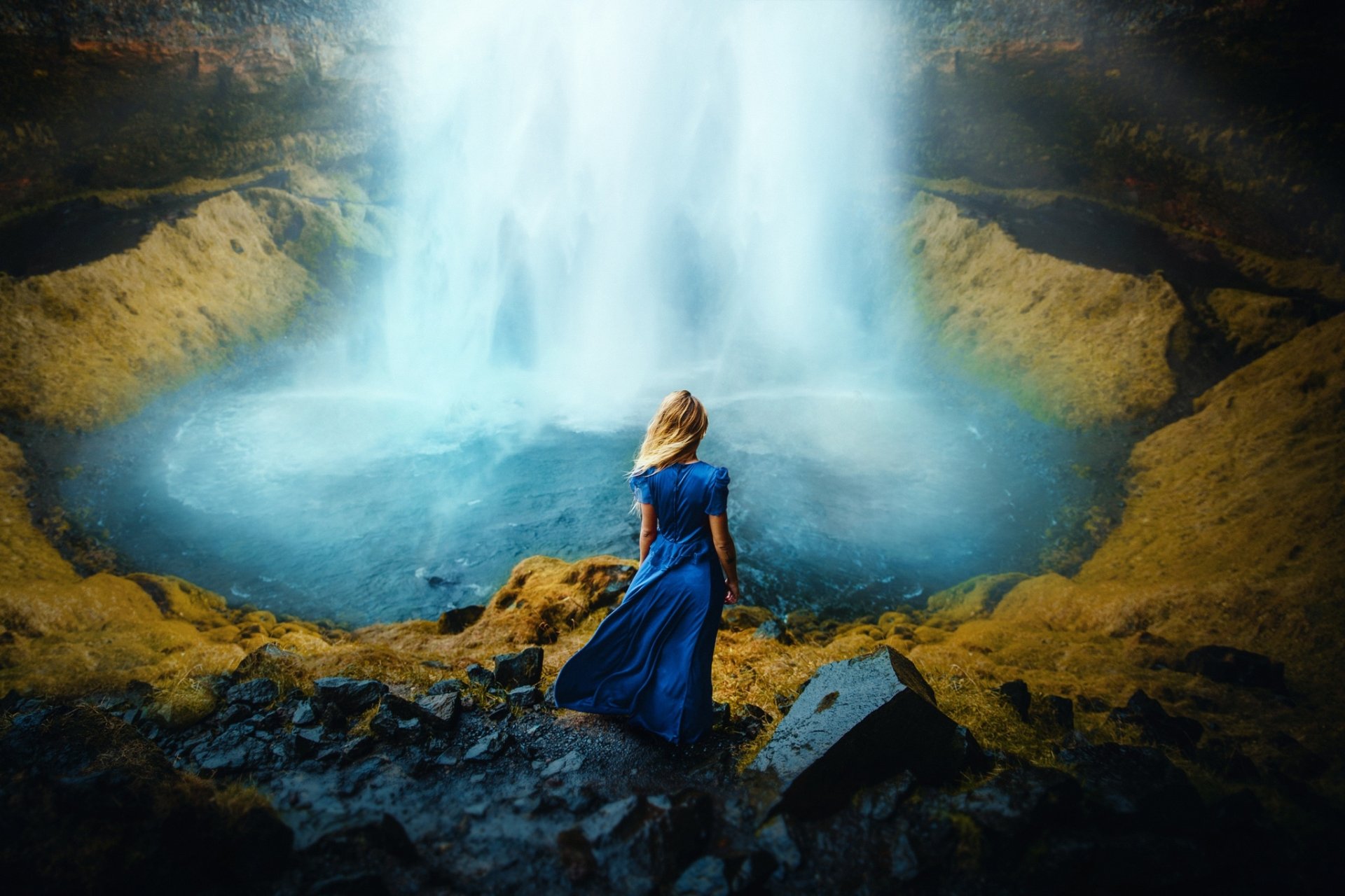 A blonde woman in a flowing blue dress stands with her back to the viewer, facing a powerful waterfall in this HD PC desktop wallpaper background.