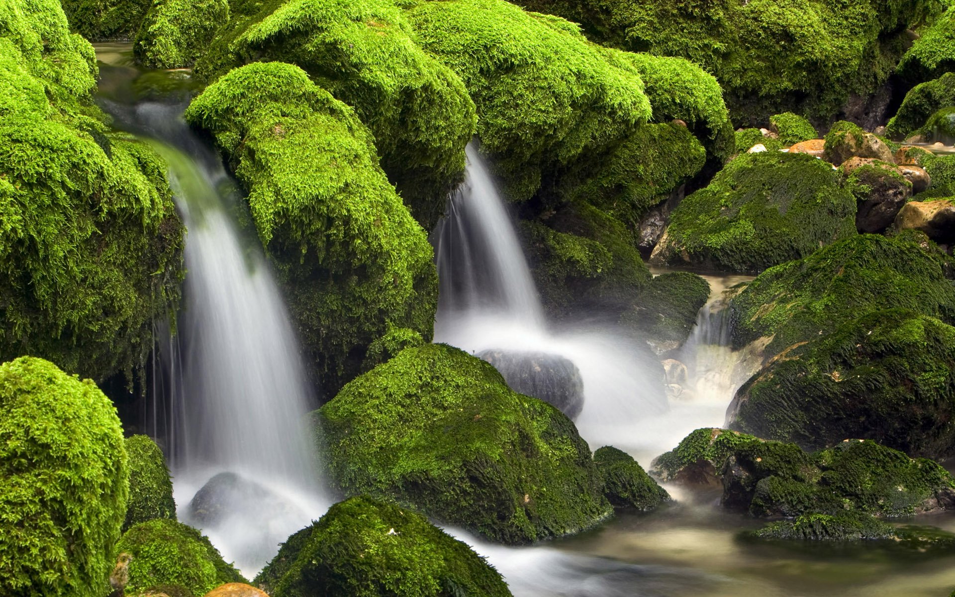 HD desktop wallpaper showcasing a serene nature scene with cascading waterfalls flowing over lush green moss-covered rocks.