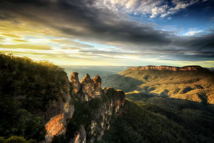 Sunlit view of the Three Sisters rock formation and lush forest cliffs in the Blue Mountains, Australia, under a dramatic sky with clouds at sunset.