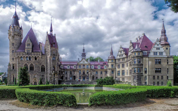 HD wallpaper of Moszna Castle in Poland, showcasing its grand architecture and surrounding garden under a partly cloudy sky.