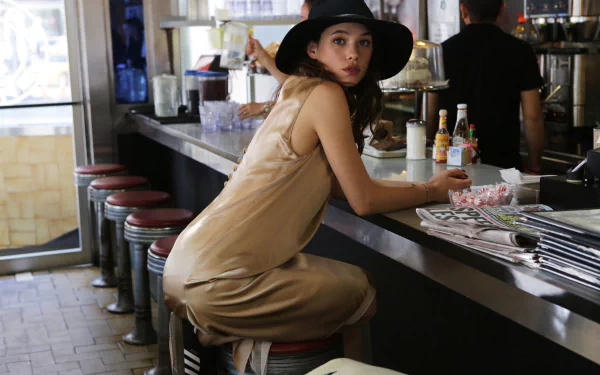 French actress Astrid Bergès-Frisbey, a brunette woman wearing a black hat and beige dress, sits at a diner counter in this HD PC desktop wallpaper.