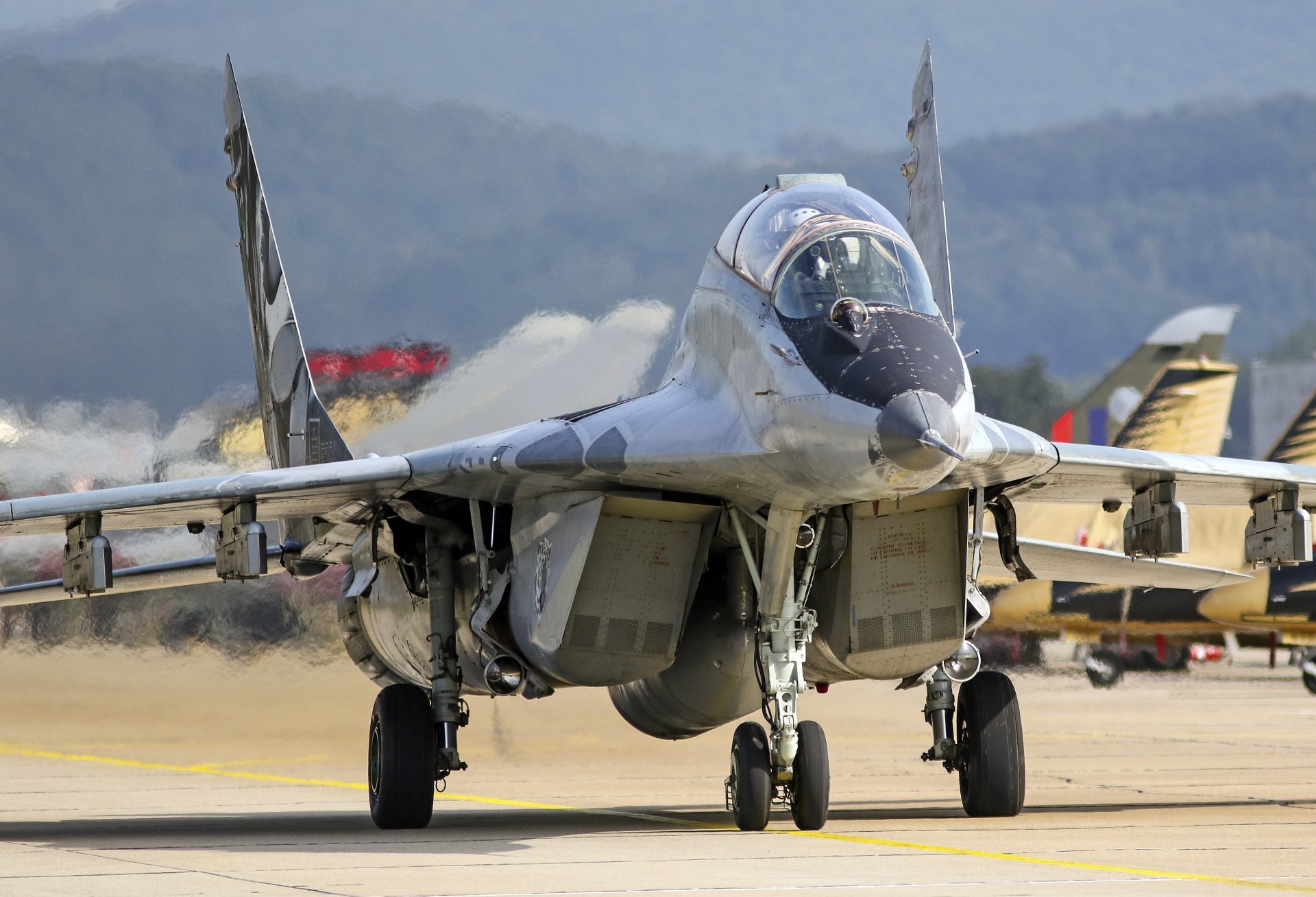 A Mikoyan MiG-29 jet fighter warplane on the runway, captured in sharp 4K Ultra HD detail against a blurred background of mountains and other aircraft.