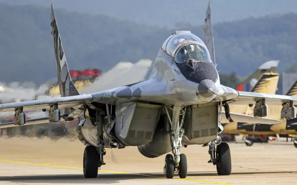 A Mikoyan MiG-29 jet fighter warplane on the runway, captured in sharp 4K Ultra HD detail against a blurred background of mountains and other aircraft.