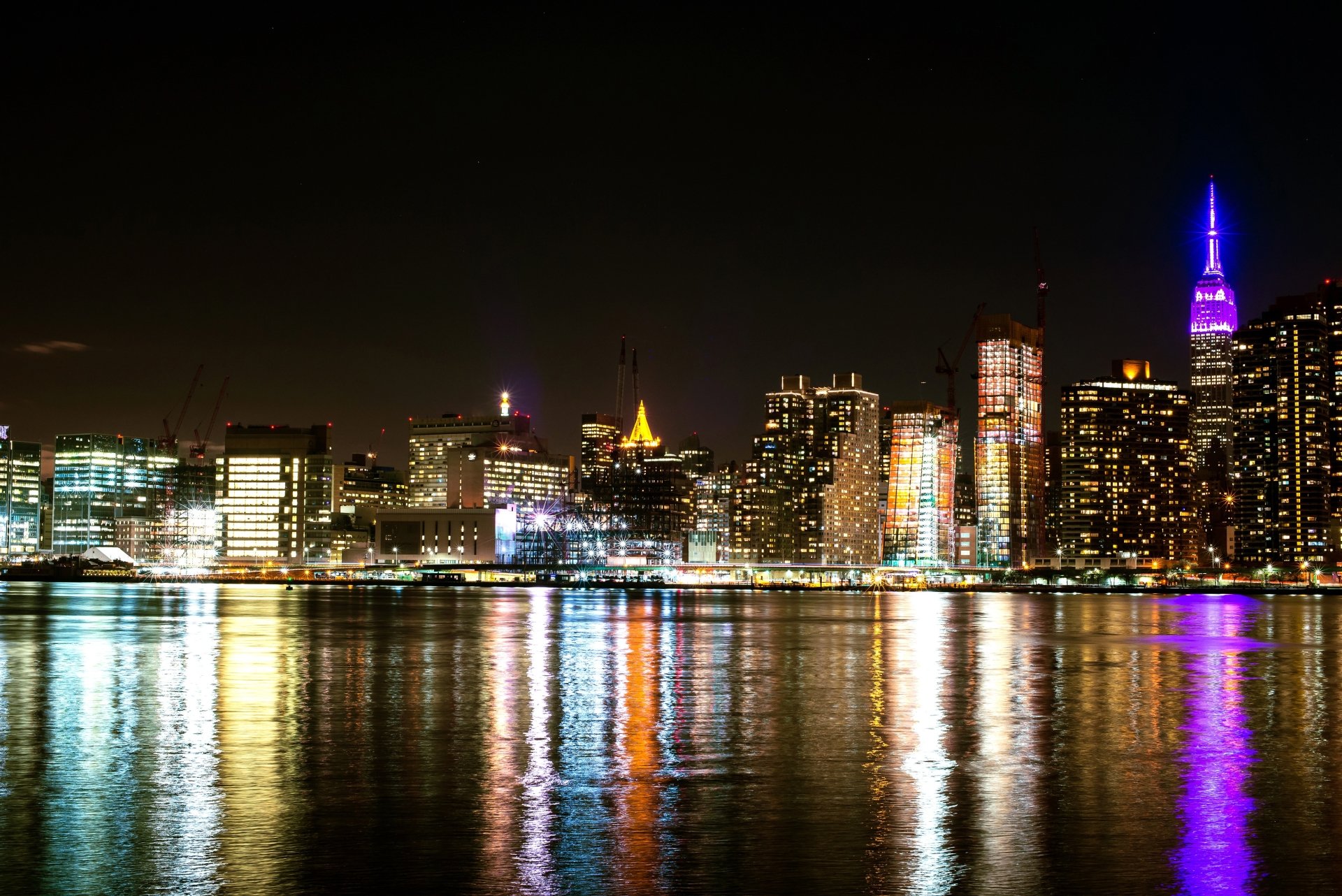 4K Ultra HD wallpaper of New York City skyscrapers at night, featuring bright building lights and colorful reflections on the water in this iconic man-made urban skyline.