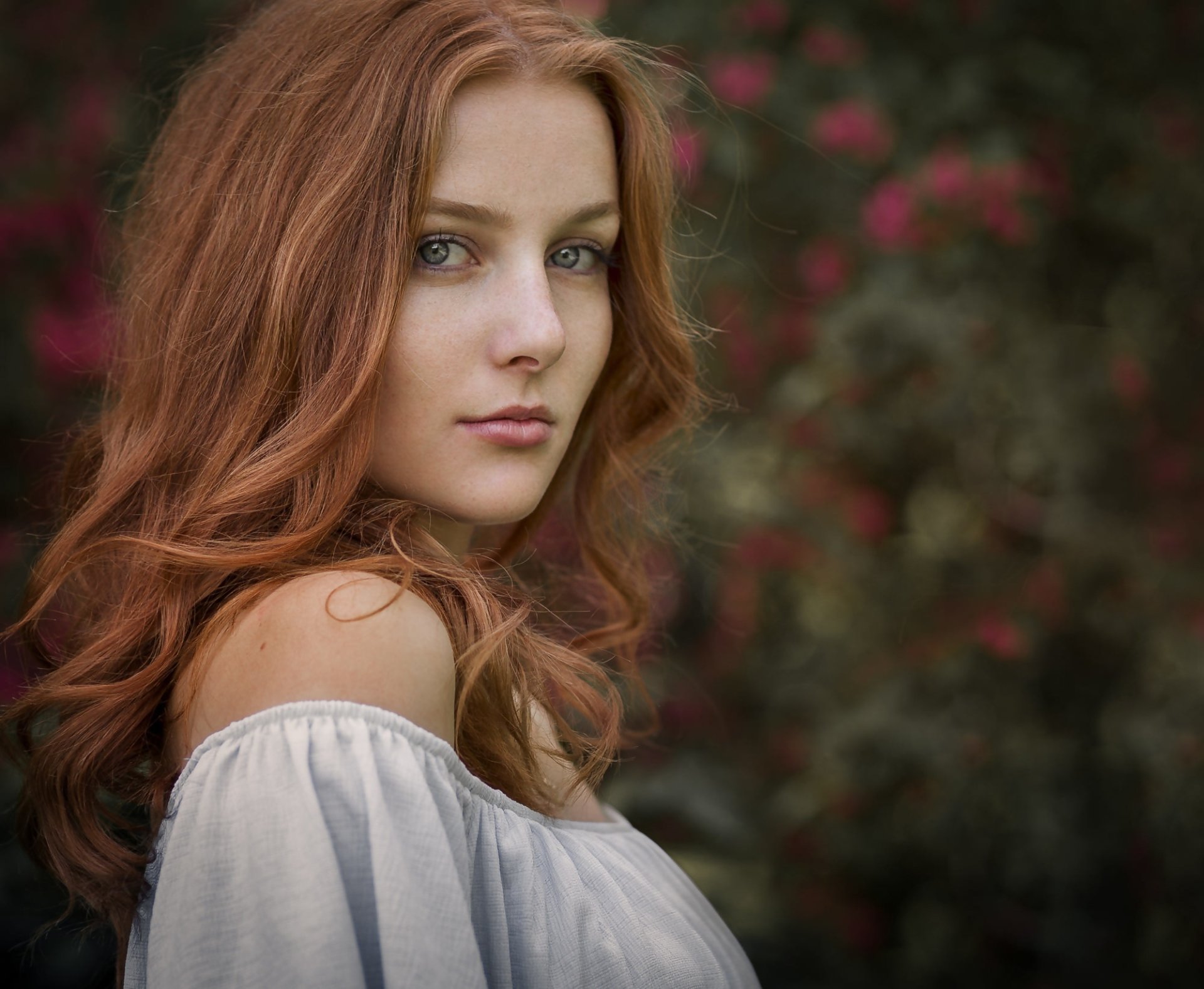 A redheaded woman with blue eyes gazes at the camera, set against a blurred floral background. This HD wallpaper captures her serene expression with a soft depth of field.