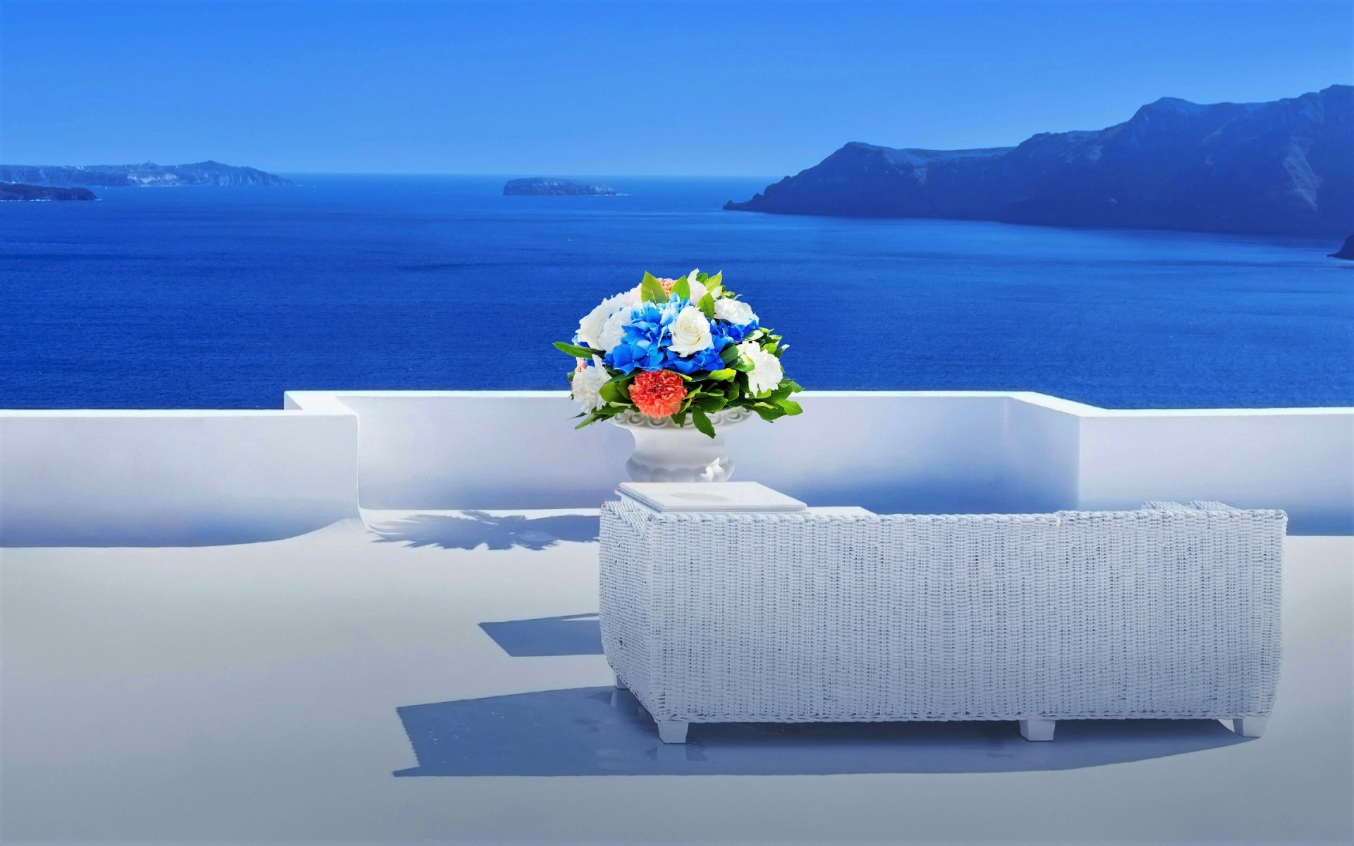 A white lounge couch and flower arrangement on a Santorini balcony overlooking the deep blue sea and horizon, with mountains in the distance under a clear sky.