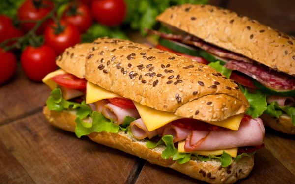 A 4K Ultra HD PC desktop wallpaper featuring two fresh sandwiches with lettuce, cheese, ham, and salami on seeded sub rolls, accompanied by cherry tomatoes on the vine.