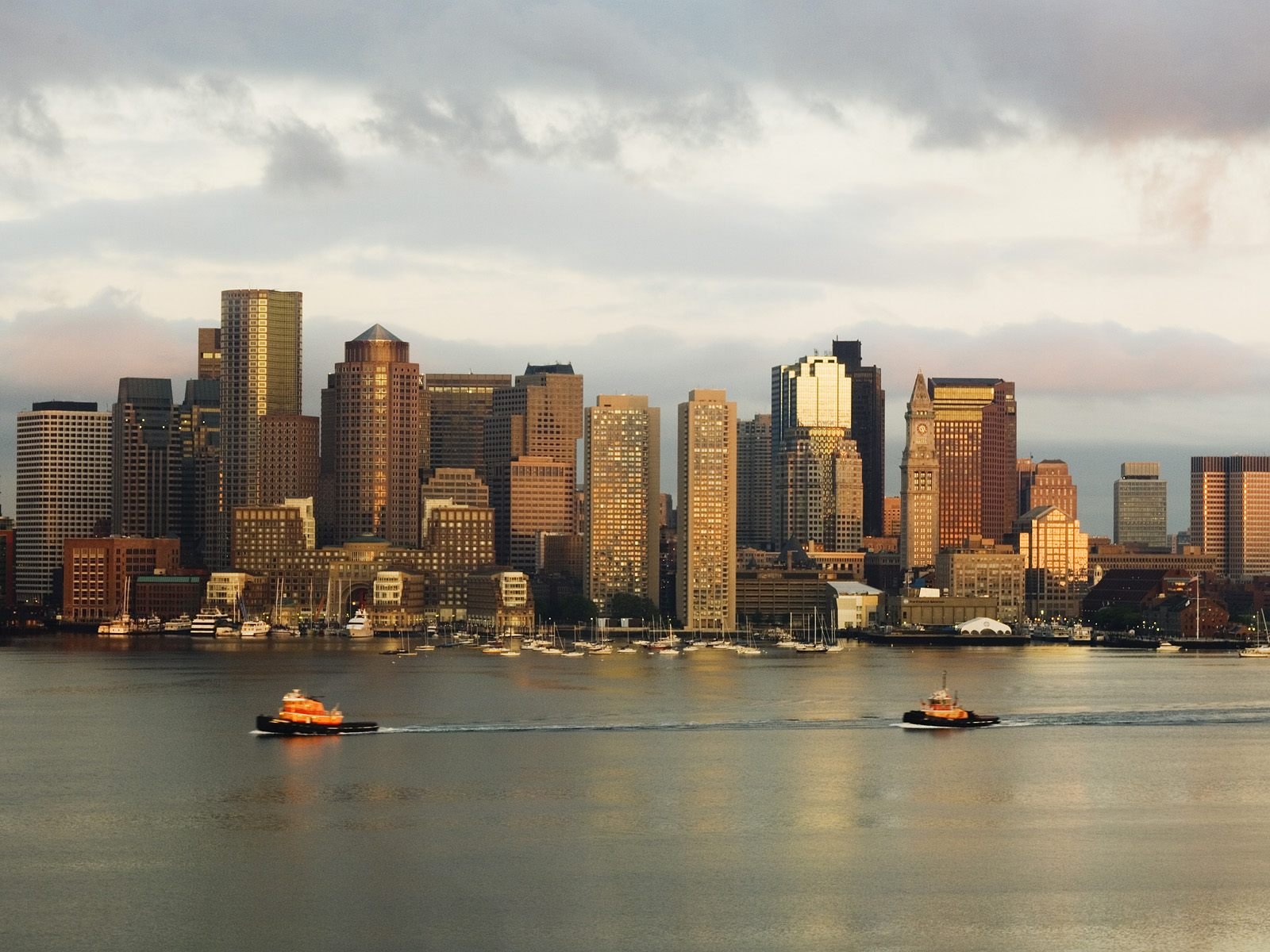 HD desktop wallpaper of Boston’s man-made skyline at sunset, featuring illuminated skyscrapers and boats crossing the calm harbor waters.