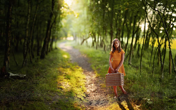 Sunlit forest path with shallow depth of field; a brunette child in an orange dress holds a wicker basket on the trail — HD photography desktop wallpaper background.