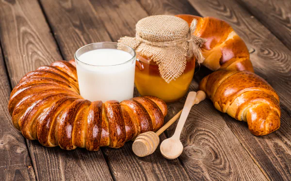 A 4K Ultra HD image of viennoiserie including croissants, a glass of milk, and a jar of honey on a wooden table, showcasing a classic breakfast spread.