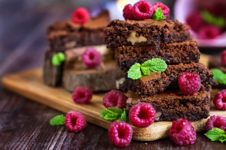 Close-up 4K Ultra HD image of rich chocolate brownies topped and surrounded by fresh raspberries and mint leaves on a wooden board, highlighting dessert and fruit textures.