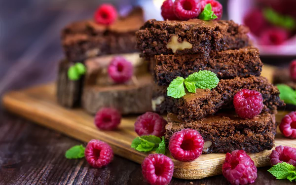 Close-up 4K Ultra HD image of rich chocolate brownies topped and surrounded by fresh raspberries and mint leaves on a wooden board, highlighting dessert and fruit textures.