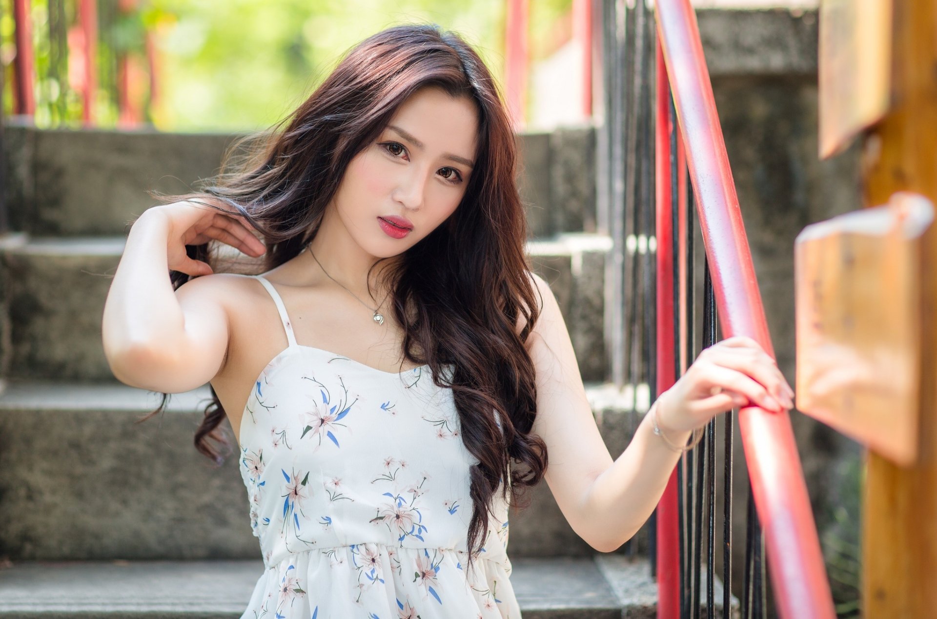 A model with long brunette hair and brown eyes stands outdoors, posing beside a red railing. The image features a depth of field effect, rendering the background slightly out of focus.