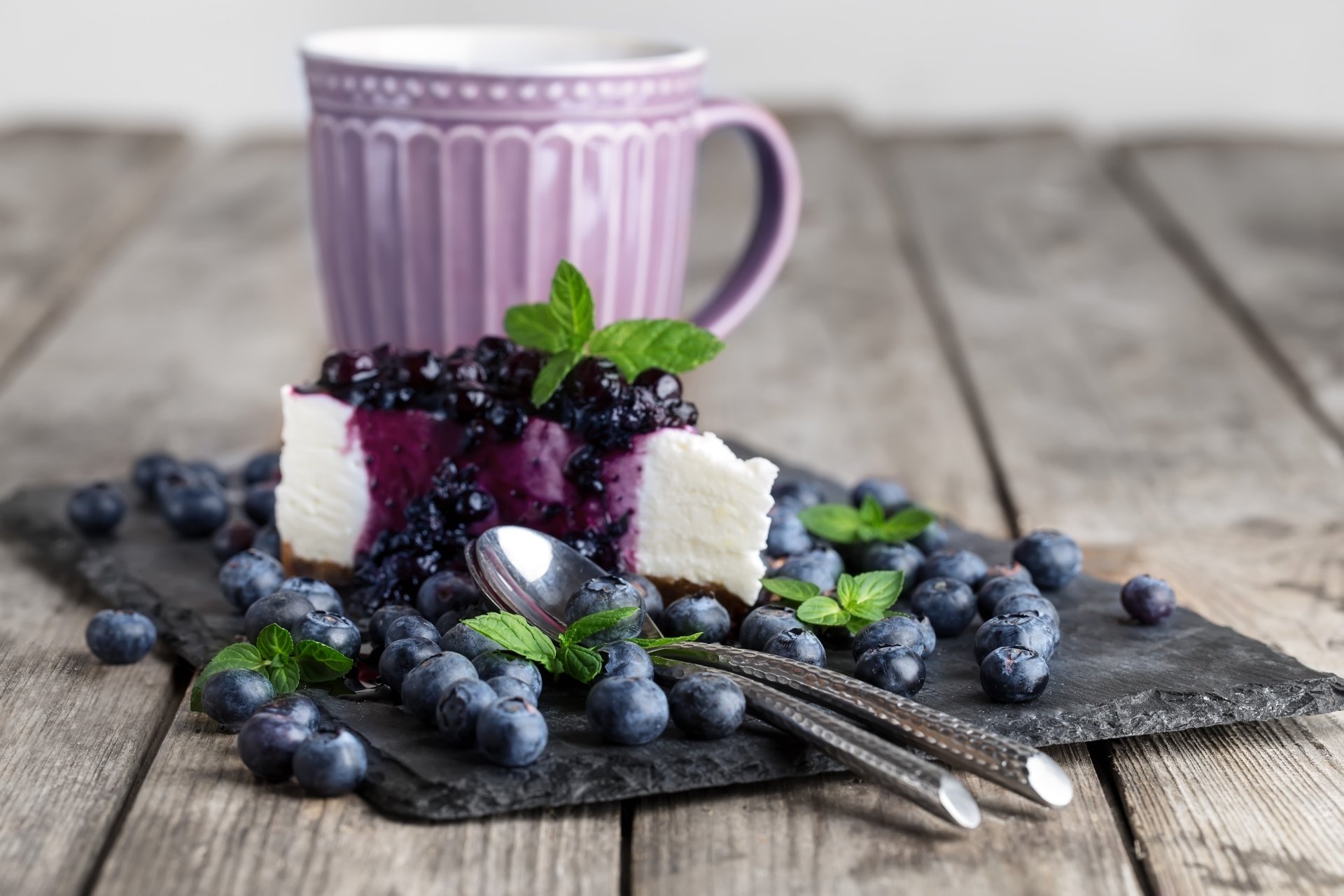 A slice of blueberry cheesecake garnished with fresh blueberries and mint leaves on a slate plate, accompanied by a purple mug, set on a rustic wooden table.
