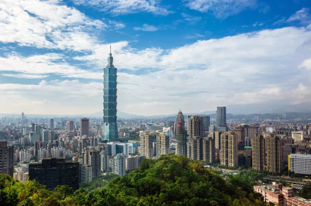 HD cityscape wallpaper featuring Taipei 101 skyscraper towering over Taipei, Taiwan, with a bright sky and scattered clouds above the urban skyline.