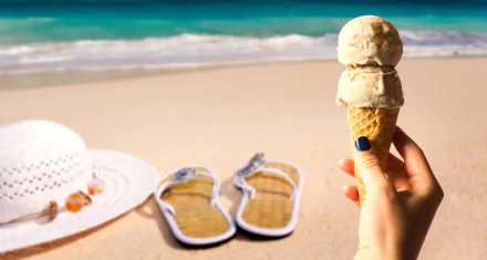 A hand holds a cone of ice cream against a sandy beach backdrop with thongs and a summer hat nearby, capturing the essence of sweet summer treats by the ocean.