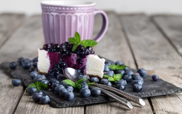 A slice of blueberry cheesecake garnished with fresh blueberries and mint leaves on a slate plate, accompanied by a purple mug, set on a rustic wooden table.