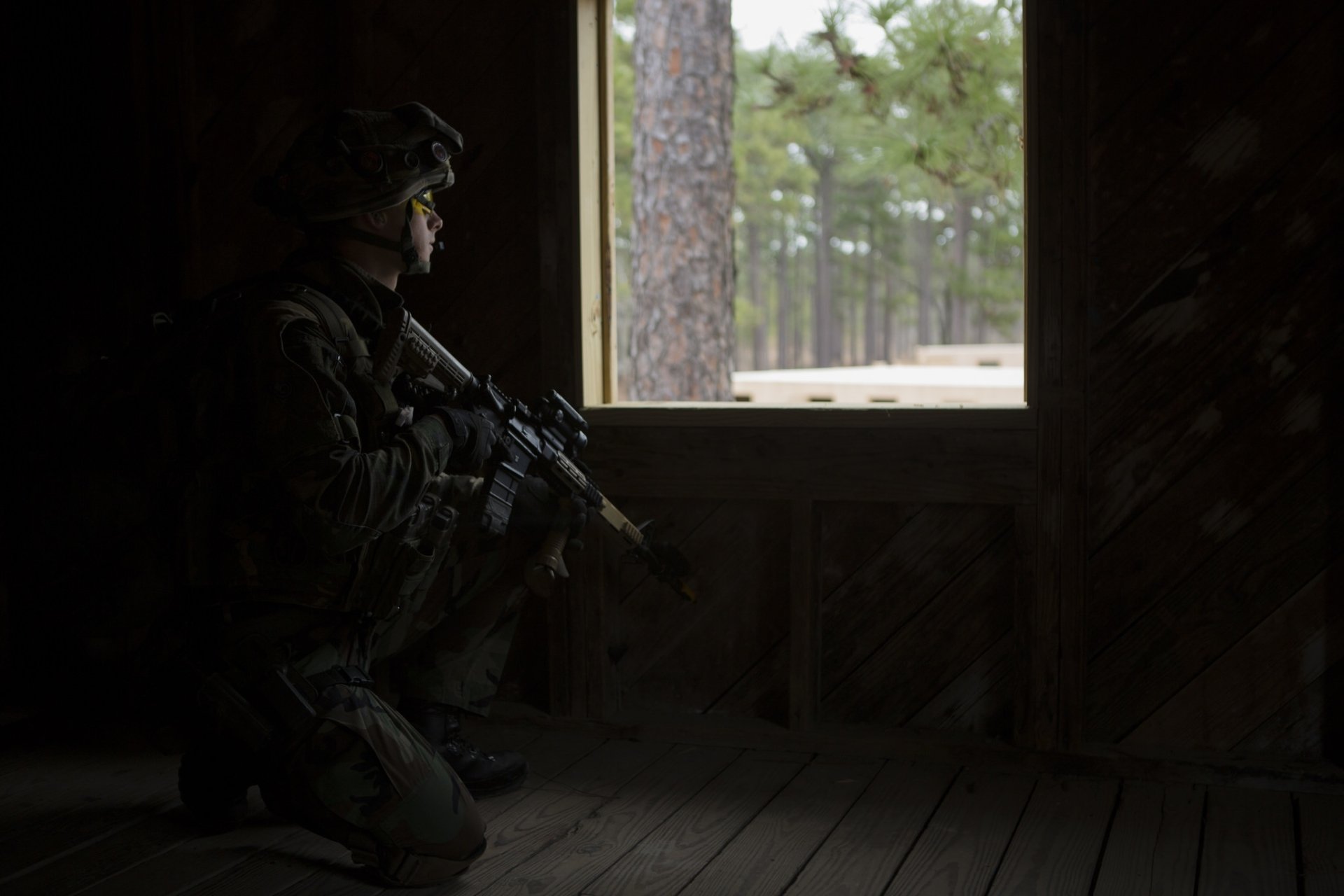 HD desktop wallpaper showing a soldier holding an M4 Carbine assault rifle inside a dark military outpost, with a forest visible through the window.