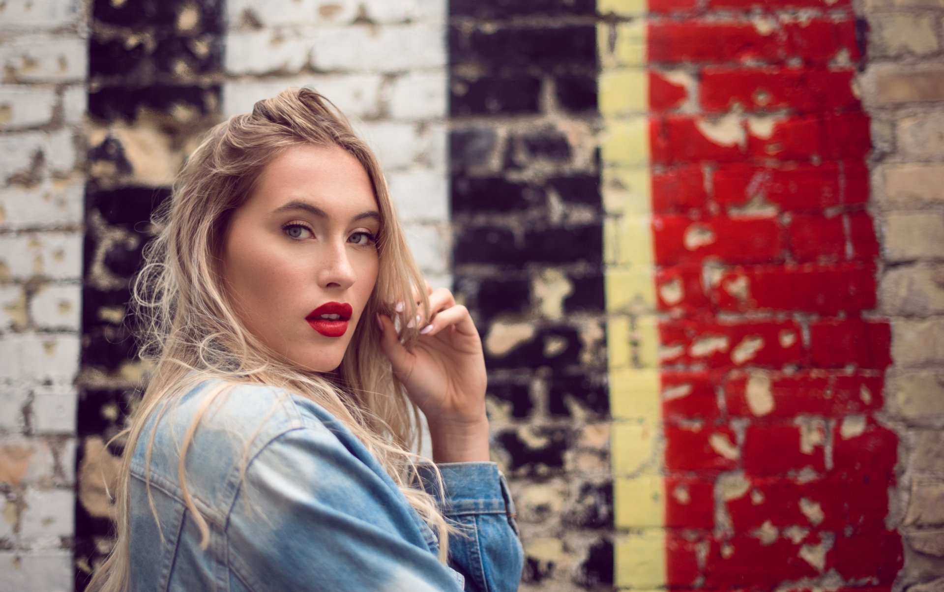 5K Ultra HD PC desktop wallpaper: blonde woman model wearing red lipstick and a denim jacket posed against a distressed, graffiti-covered brick wall background.
