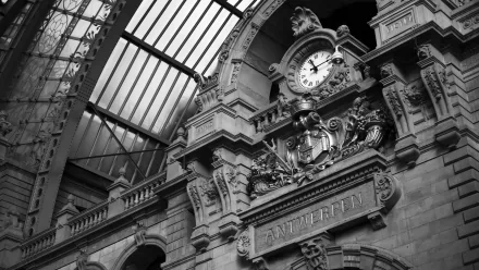 Black and white photo of an ornate concrete building interior featuring a large clock and intricate architectural shapes under a glass ceiling, captured in high definition.