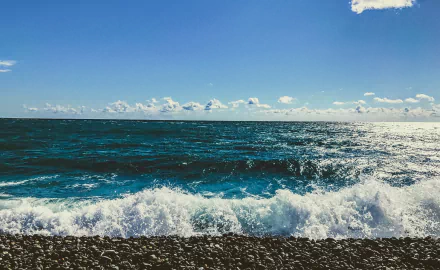 HD desktop wallpaper showing a vibrant ocean scene with waves crashing on the shore under a bright blue sky along the horizon.