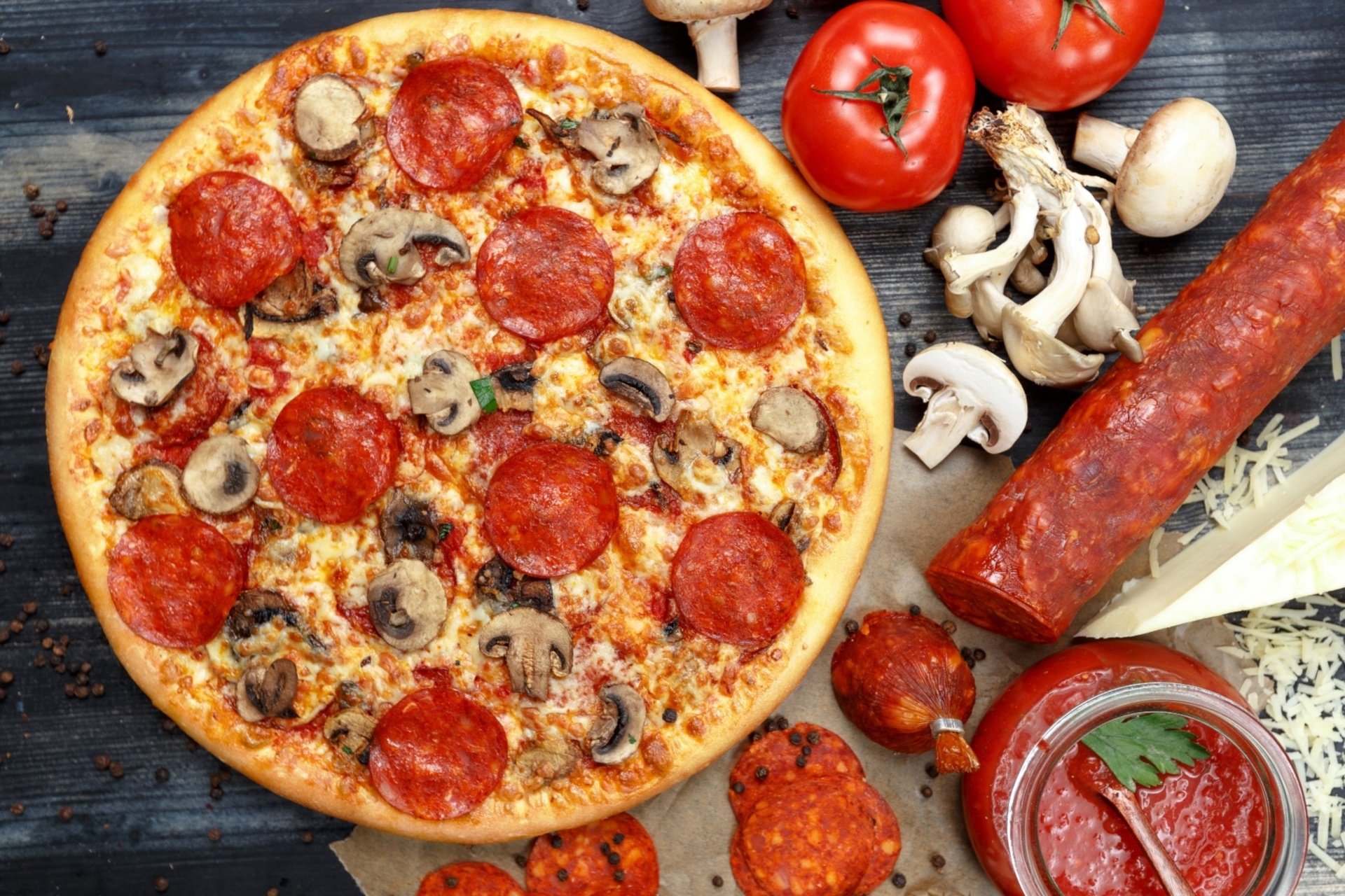 HD desktop background featuring a still life of a pepperoni and mushroom pizza surrounded by fresh tomatoes, sausage, mushrooms, and tomato sauce.