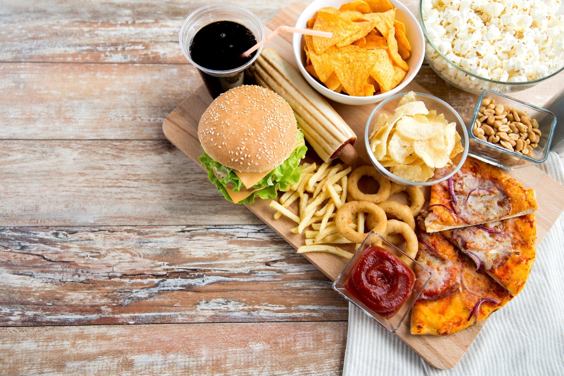 HD desktop wallpaper featuring a still life spread of fast food including a burger, pizza slices, fries, chips, popcorn, peanuts, and a soda on a wooden surface.