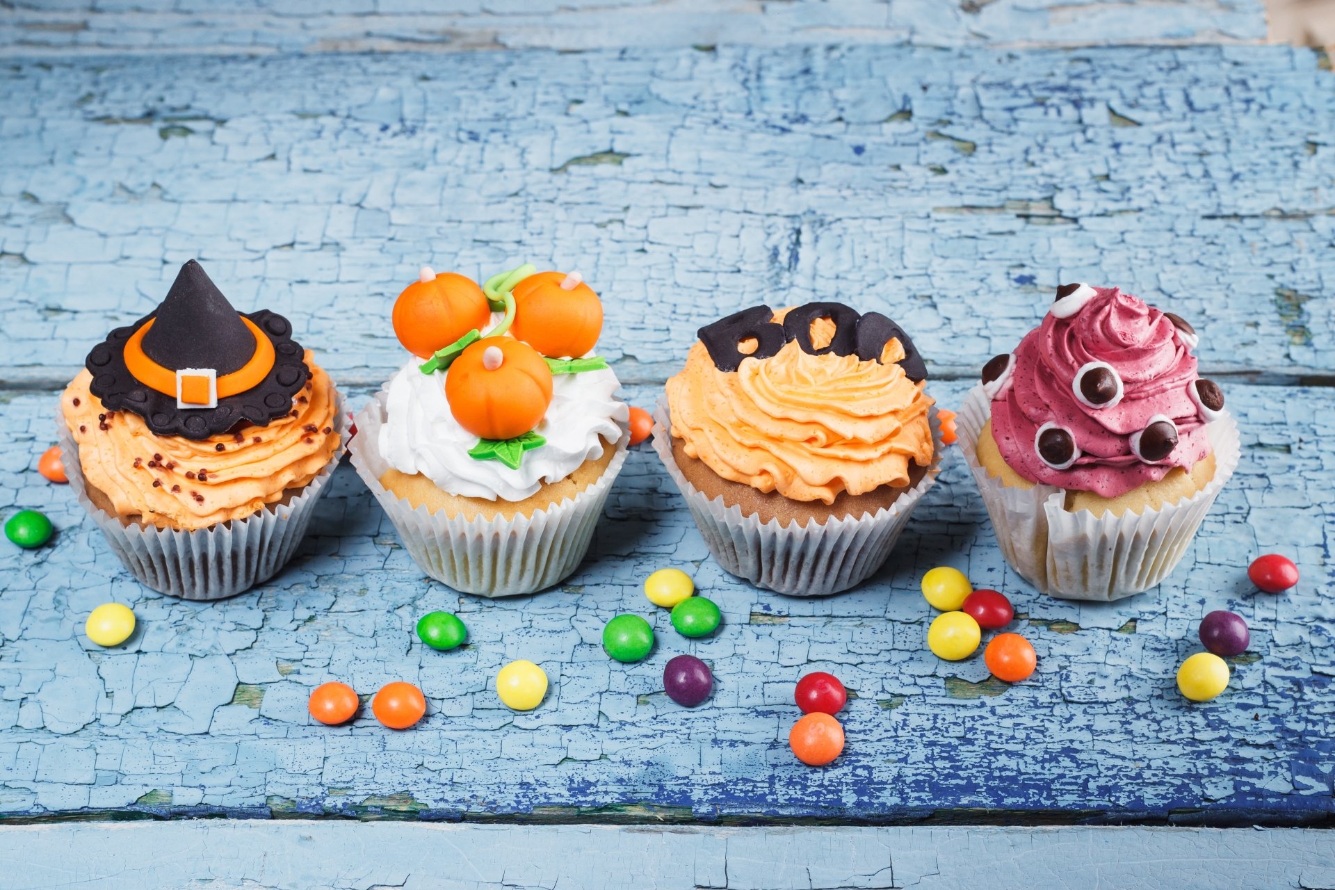 Four Halloween-themed cupcakes topped with cream, candy decorations, and colorful candy pieces scattered on a rustic blue wooden surface in 4K Ultra HD.
