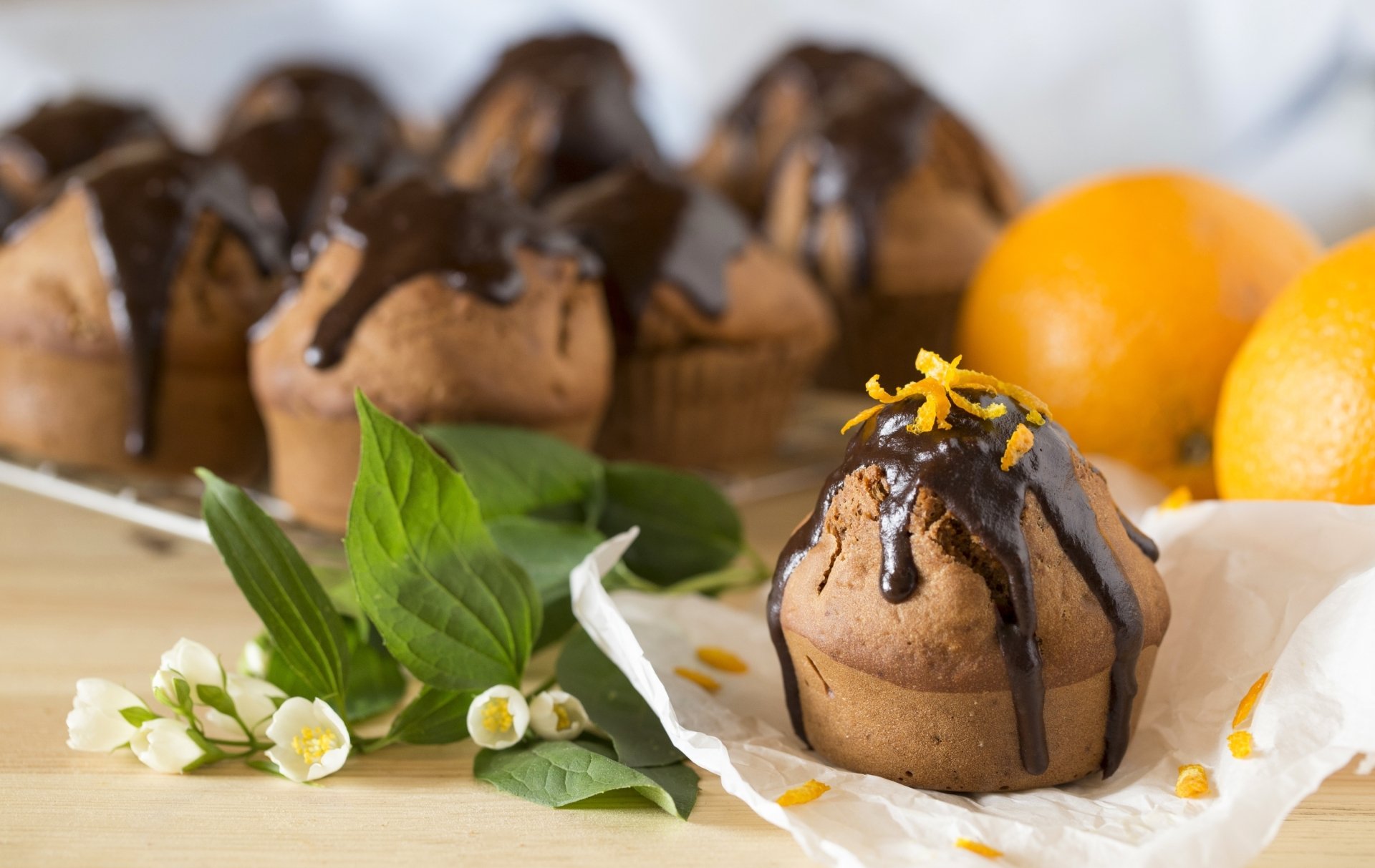 HD desktop wallpaper of a still life featuring chocolate-drizzled muffins, fresh oranges, and green leaves with small white flowers arranged on a wooden surface.
