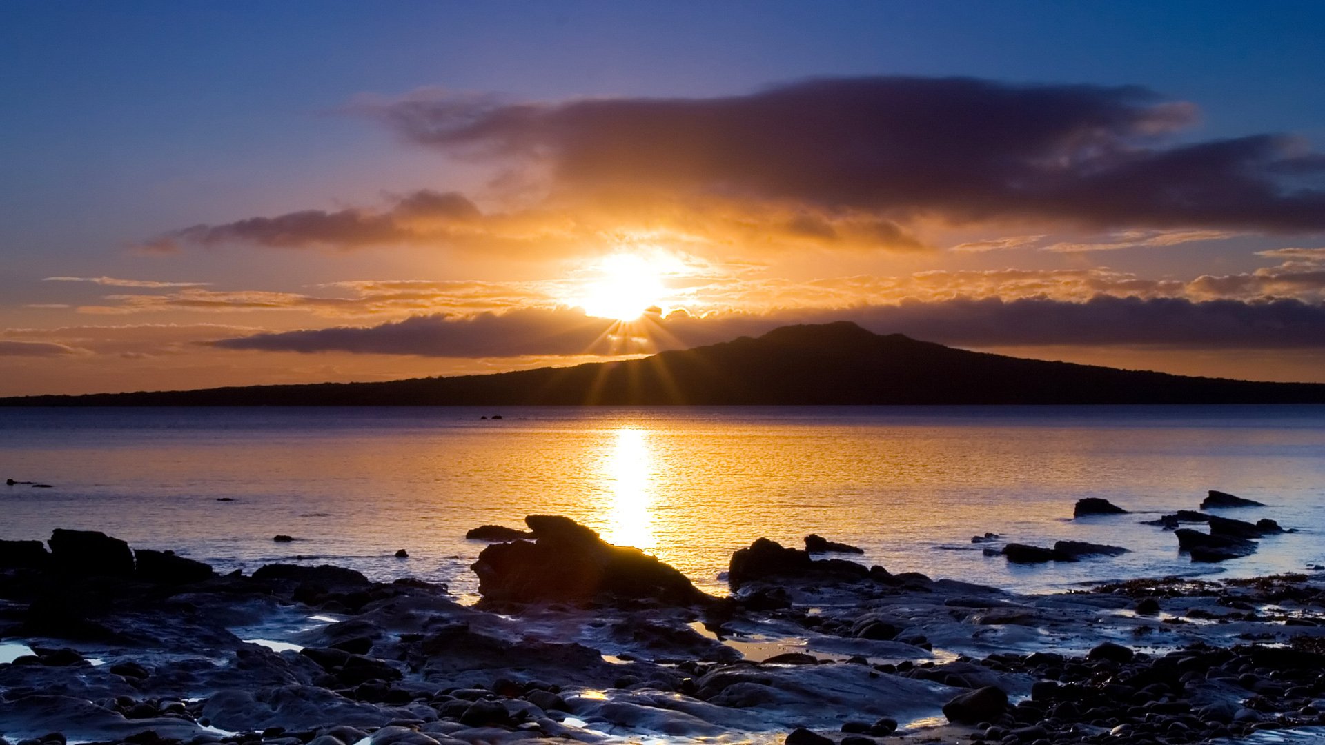 Sunrise or sunset over a mountainous shoreline with clouds reflecting on calm ocean water, capturing the beauty of wilderness in this HD landscape nature wallpaper.