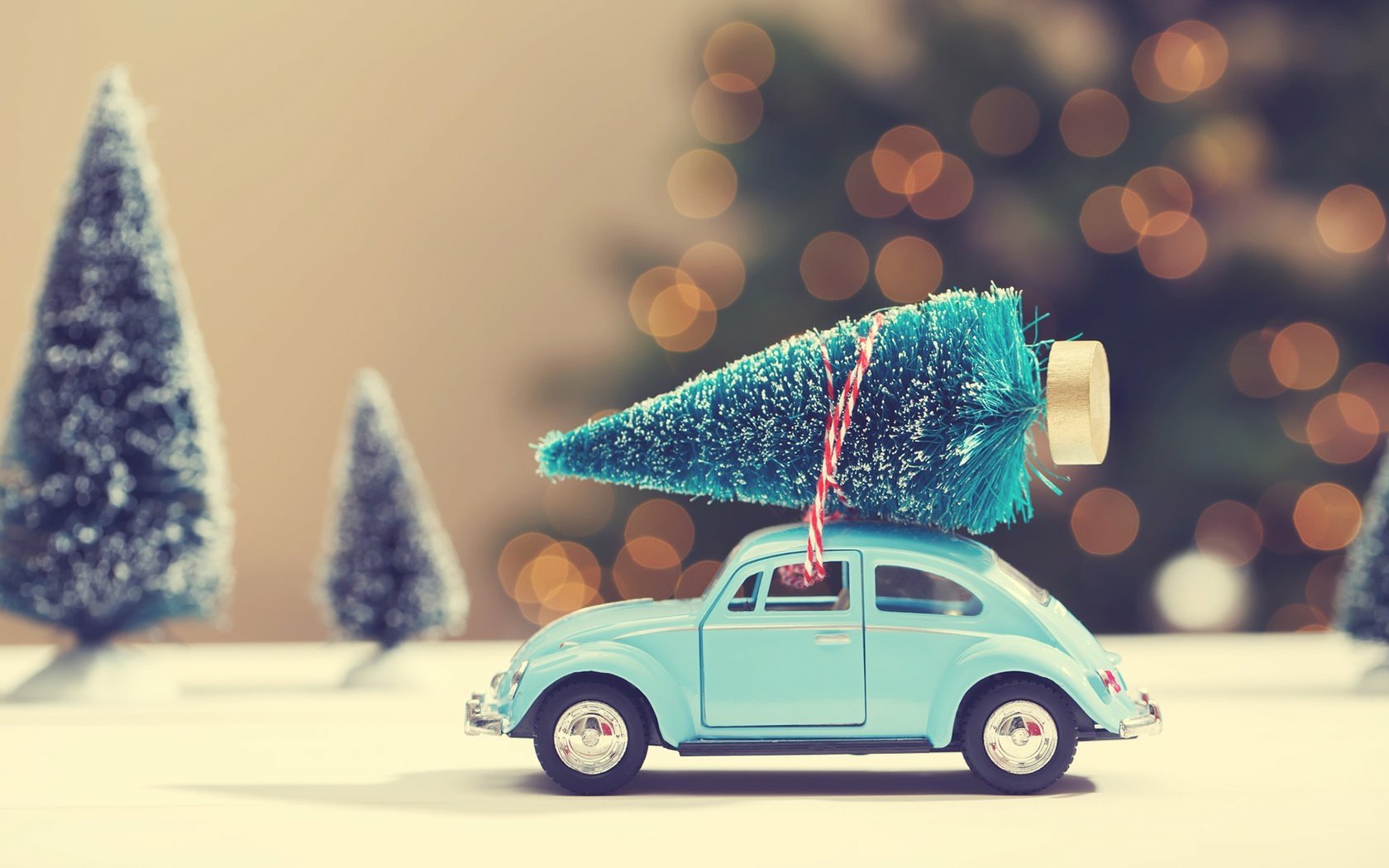A toy car carrying a miniature Christmas tree with holiday bokeh lights in the background, presented as an HD PC desktop wallpaper and background.
