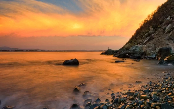 A stunning sunrise over the ocean illuminates the sky with vibrant colors, reflecting on gentle waves lapping against a pebble-strewn beach beside a hillslope.