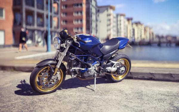 HD desktop wallpaper featuring a blue Ducati motorcycle with a shallow depth of field, parked near a waterfront with urban buildings in the background.