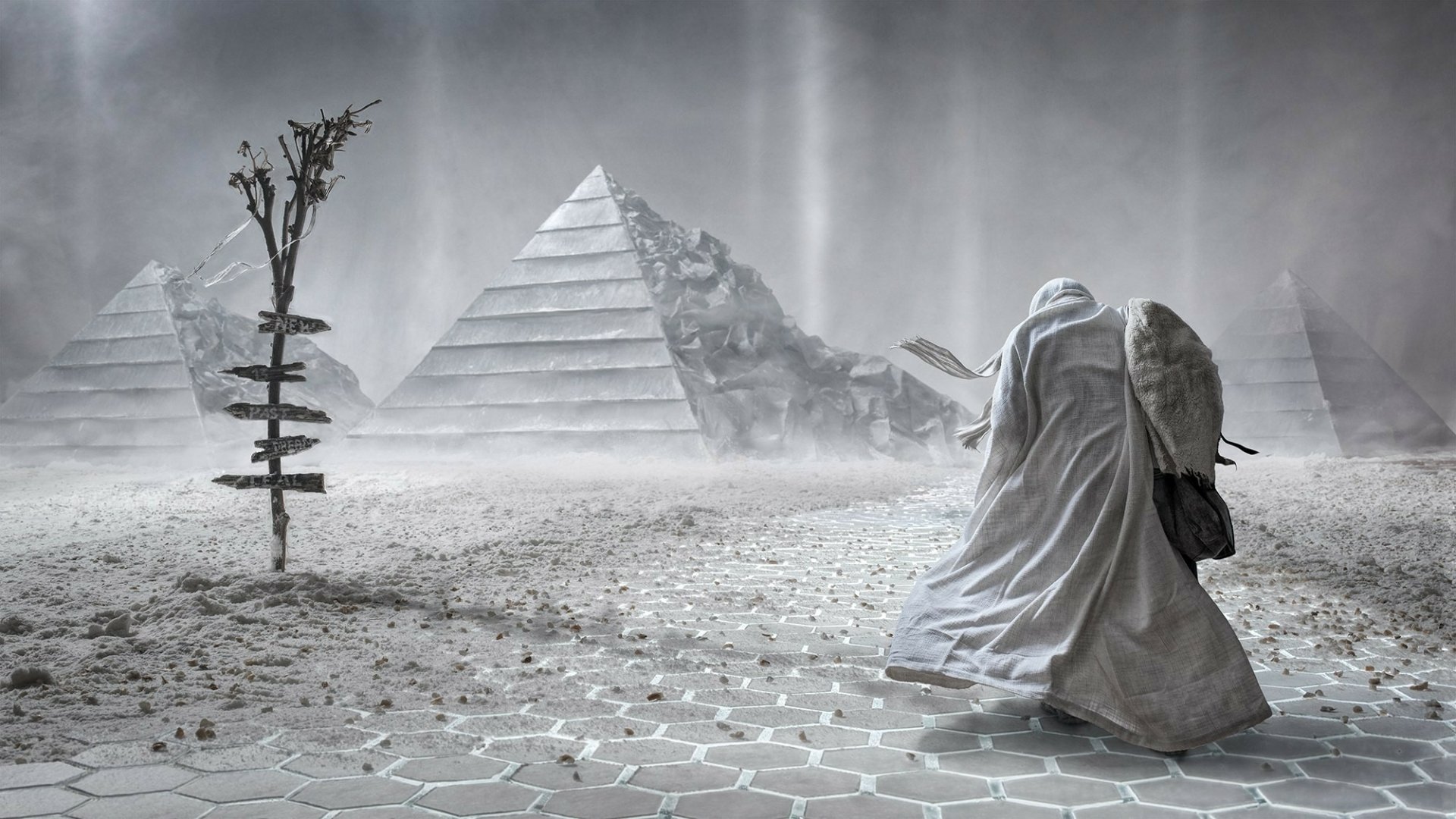 A fantasy warrior in a tattered cloak stands in a desolate landscape with pyramids in the background, draped in mist and shadows, creating an eerie HD desktop wallpaper.