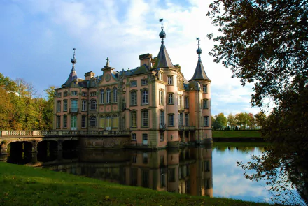 Poeke Castle, a historic man-made castle in Belgium, stands surrounded by trees and reflected in a calm lake under a partly cloudy sky.