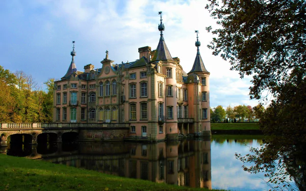 Poeke Castle, a historic man-made castle in Belgium, stands surrounded by trees and reflected in a calm lake under a partly cloudy sky.