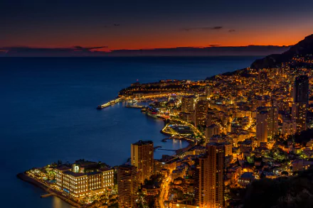 Nighttime view of Monaco's city skyline with illuminated skyscrapers along the coastline, captured in high definition against a deep blue horizon.