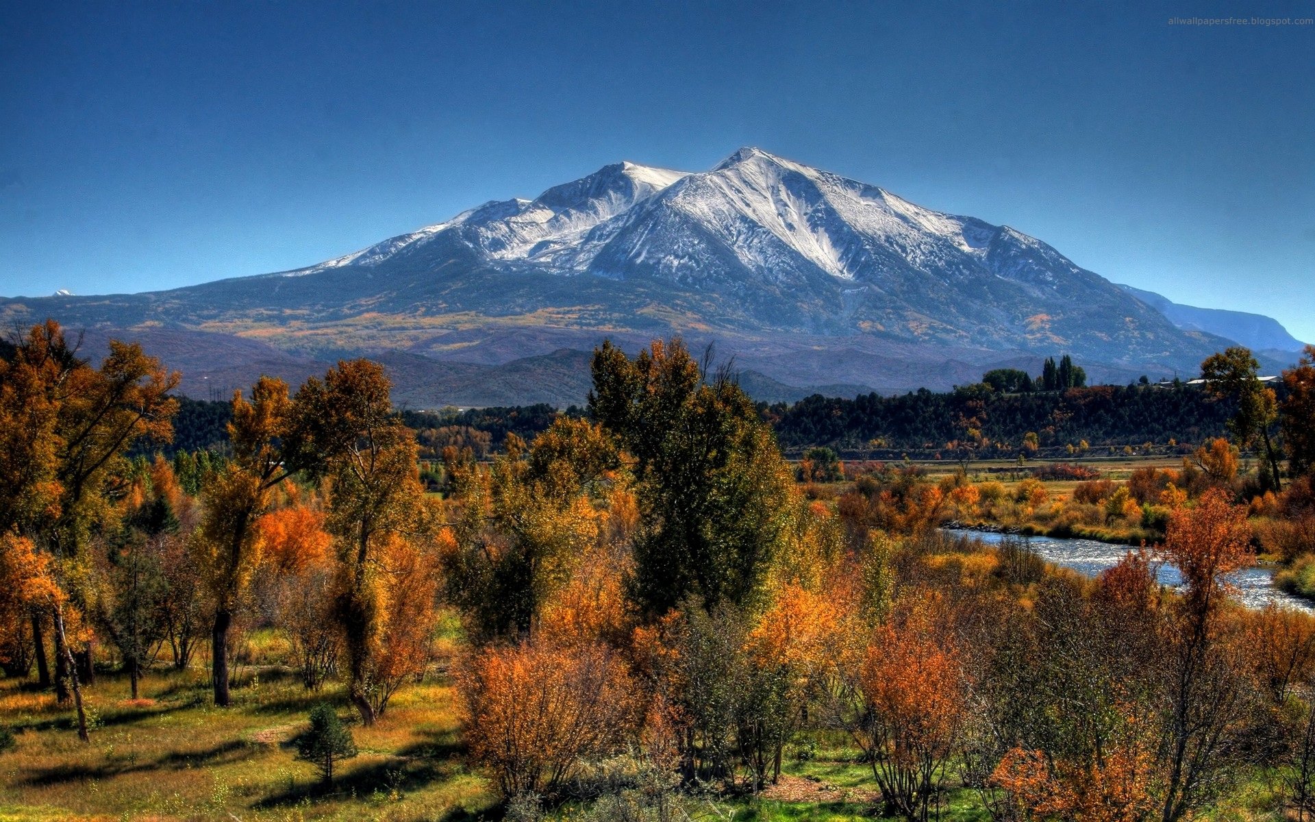 Snow-capped mountain towers over a winding river, surrounded by autumn-colored trees, grassy fields, and a clear blue sky in a vibrant nature landscape.