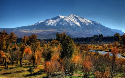 Snow-capped mountain towers over a winding river, surrounded by autumn-colored trees, grassy fields, and a clear blue sky in a vibrant nature landscape.