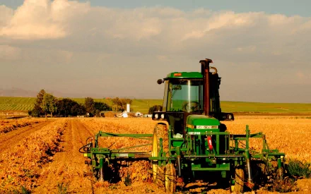 A John Deere vehicle working in a sunlit field, surrounded by cultivated land and distant trees, creates a striking HD desktop wallpaper and background.