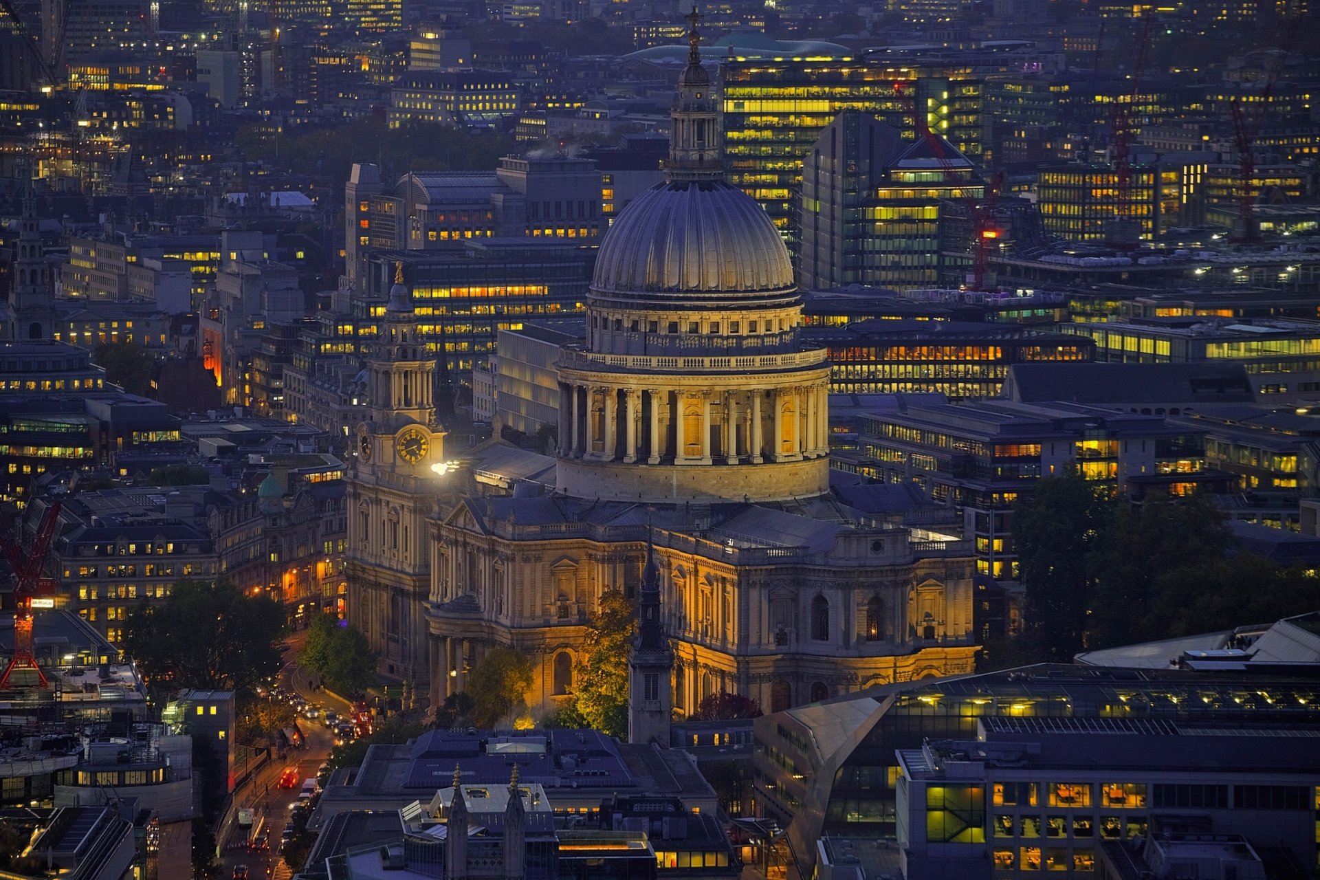 Night view of St Paul's Cathedral dome illuminated amidst the cityscape of London, England, showcasing the religious landmark surrounded by urban buildings.