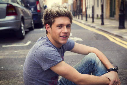 HD wallpaper featuring a smiling man in a grey t-shirt sitting on a city street, for Niall Horan fans' desktop backgrounds.