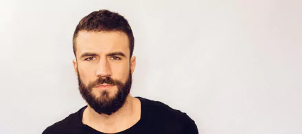 HD desktop wallpaper featuring a portrait of a bearded man in a black shirt against a light background.