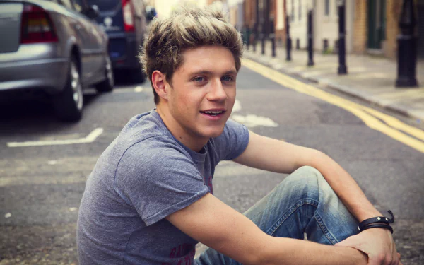 HD wallpaper featuring a smiling man in a grey t-shirt sitting on a city street, for Niall Horan fans' desktop backgrounds.
