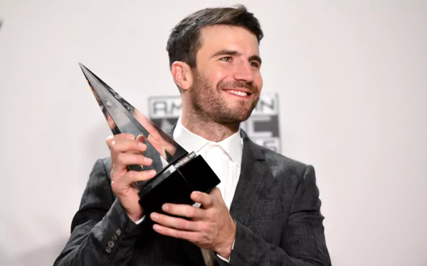 Sam Hunt smiles while holding a trophy, dressed in a stylish suit against a simple background. This HD wallpaper captures his success in the music industry.