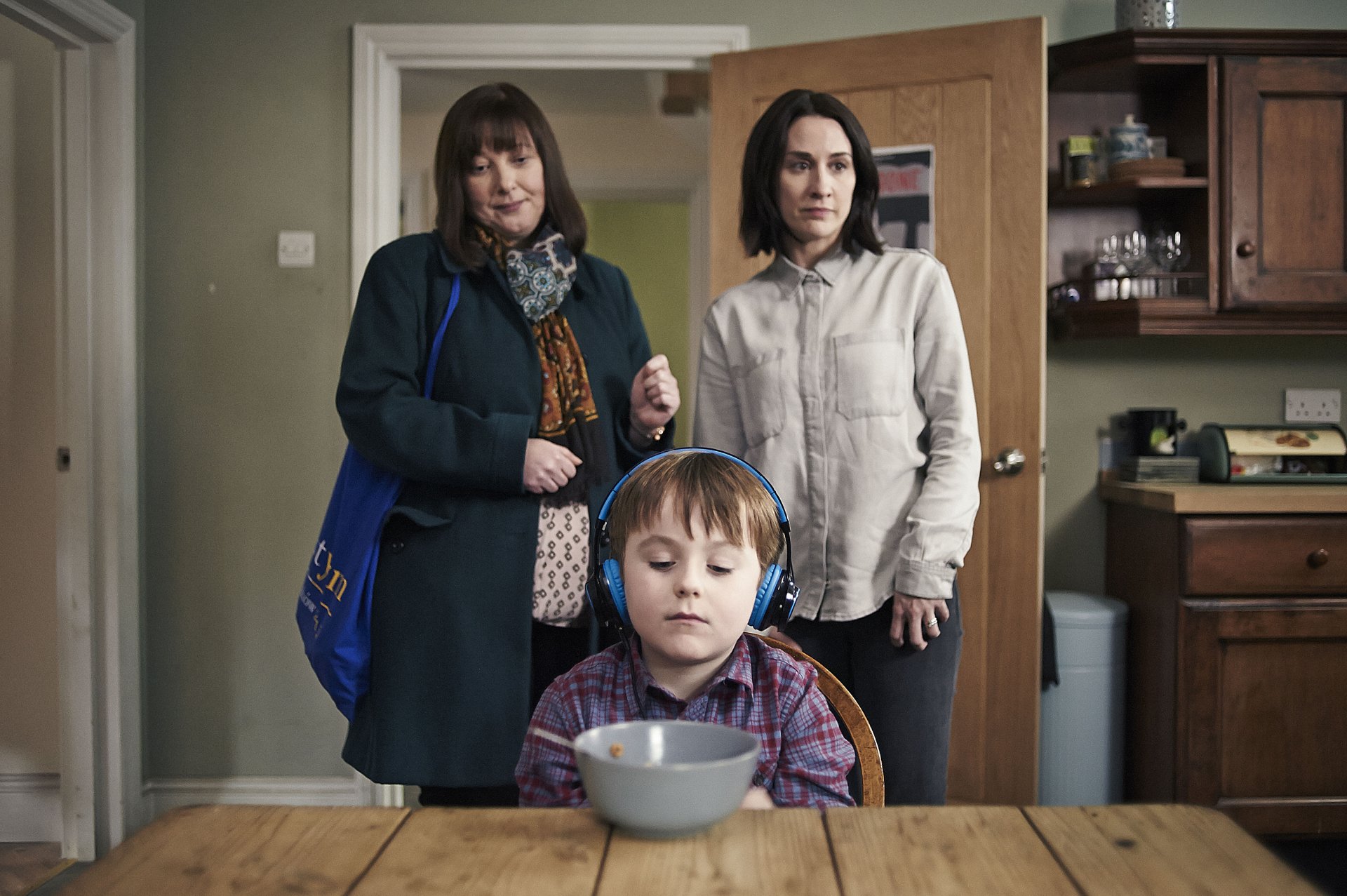 HD desktop wallpaper featuring characters from The A Word with a child wearing headphones at a table and two women standing behind.