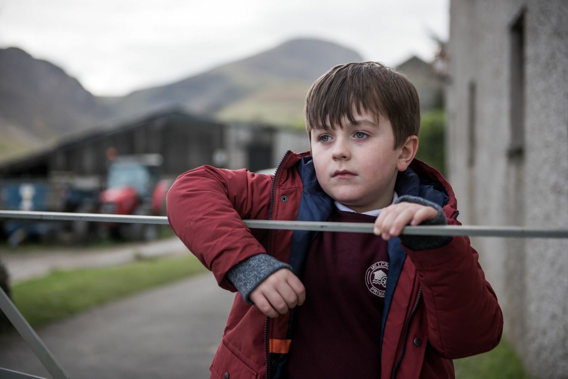 HD desktop wallpaper from The A Word featuring a thoughtful young boy in a red coat with scenic hills in the background.