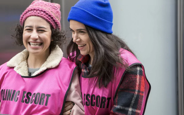 HD wallpaper featuring two smiling characters from Broad City, wearing colorful hats and pink vests labeled NANNYING ESCORT.
