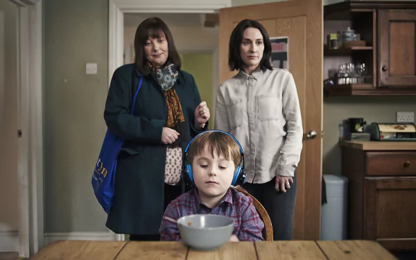 HD desktop wallpaper featuring characters from The A Word with a child wearing headphones at a table and two women standing behind.