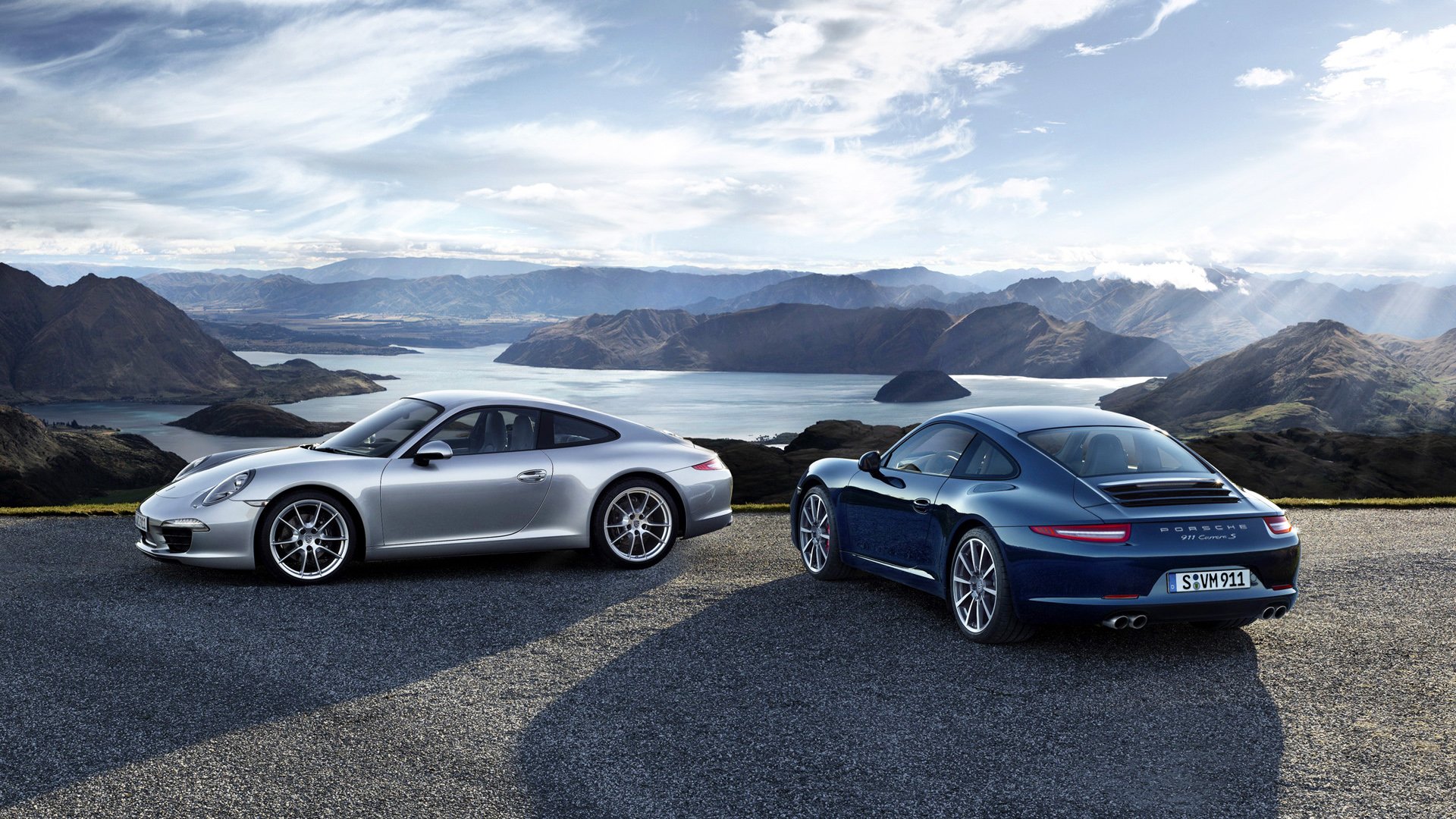 HD PC desktop wallpaper showing two Porsche 911 Carrera vehicles (silver and blue) parked on a mountain overlook above a lake under a dramatic cloudy sky.